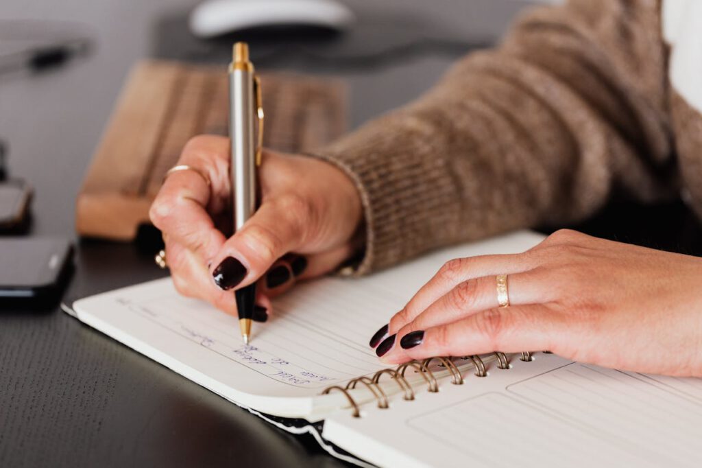 zipwp-image-4476376 Crop unrecognizable female with stylish manicure sitting at black desk with keyboard and smartphone and taking notes with silver pen in notepad